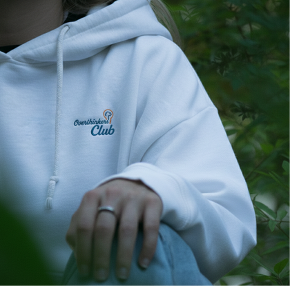Person wearing a white hoodie with 'Overthinkers Club' logo, sitting outdoors.