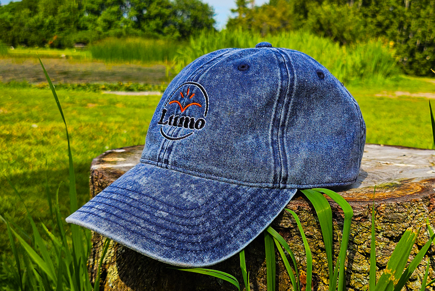 Blue denim cap with a Lumo logo on a wooden stump in a grassy field